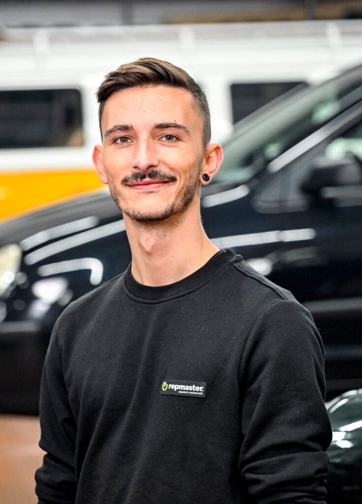Person in schwarzer Arbeitskleidung mit „repmaster“-Logo, im Hintergrund ein gelber VW-Bus und ein schwarzes Fahrzeug.