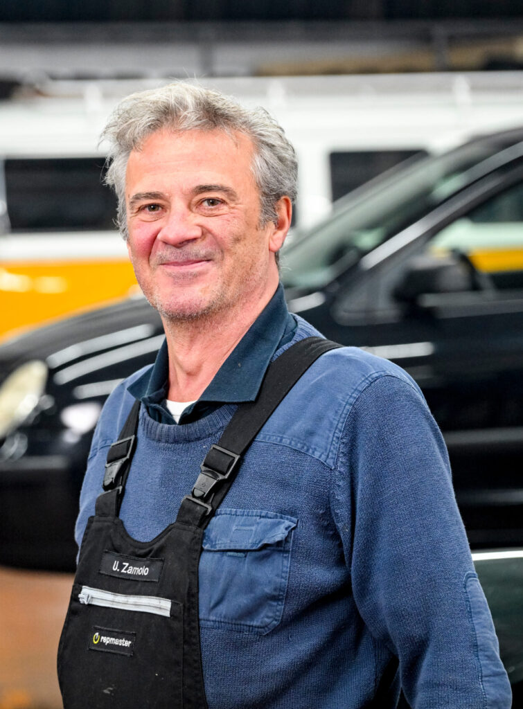 Mechaniker in Arbeitskleidung, im Hintergrund ein gelber VW-Bus und ein schwarzes Fahrzeug.
