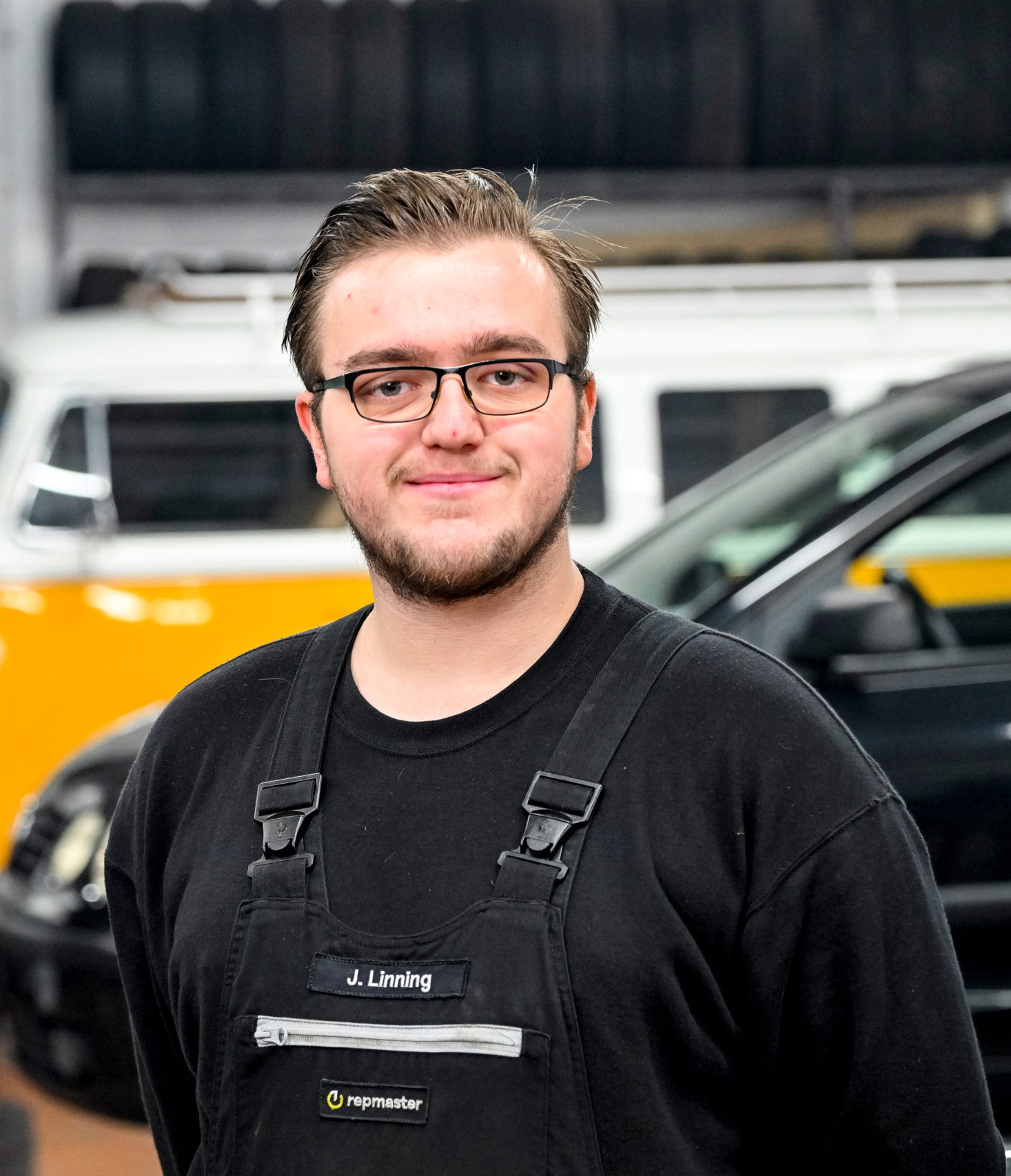 Ein Werkstattmitarbeiter in schwarzer Arbeitskleidung mit Namensschild steht in einer Garage, im Hintergrund sind ein gelber Oldtimer-Bus und ein schwarzes Fahrzeug sichtbar.